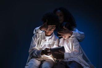 Mãe acompanha duas crianças enquanto usam um tablet em ambiente doméstico.